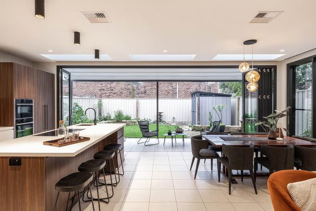 A bright, open-plan contemporary kitchen and dining area with a large island, dark timber cabinetry, integrated appliances, and bar seating. The space opens through extensive glazing and sliding doors to a paved outdoor courtyard, while a dining table, sculptural pendant lights, and indoor plants create a connected entertaining zone.