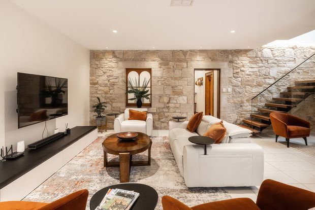 A refined open-plan living room with white sectional sofas, warm leather accent chairs, layered round coffee tables, and a wall-mounted television on sleek built-in joinery. The room features a prominent rough natural-stone feature wall with recessed artwork and a doorway, a tiled floor, recessed ceiling lighting, and an open-riser timber-and-metal staircase. The palette is warm and neutral with contemporary furnishings and rustic architectural detailing.