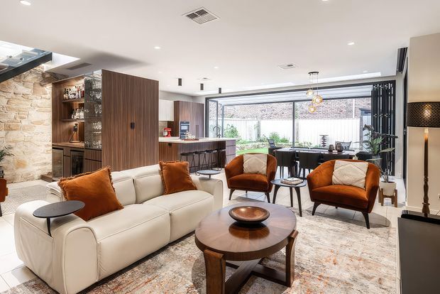 A bright, open-plan contemporary living room with a large cream sectional sofa, rust-orange accent chairs and cushions, and a round timber coffee table. The space includes a dining area and kitchen beyond, extensive glazing overlooking a courtyard, built-in timber cabinetry and shelving, a natural stone feature wall, recessed ceiling lighting, and a patterned area rug.