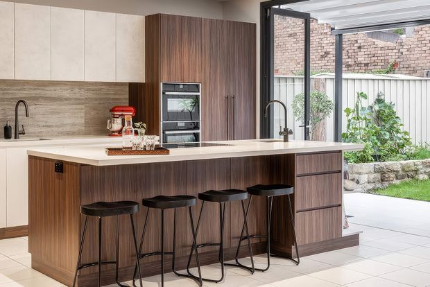 A contemporary kitchen centred on a large island with seating for four. The kitchen features dark timber-look cabinetry, pale upper cabinets, a light stone island benchtop, integrated appliances, a textured stone-look splashback, and black-framed glazing opening toward a landscaped courtyard.