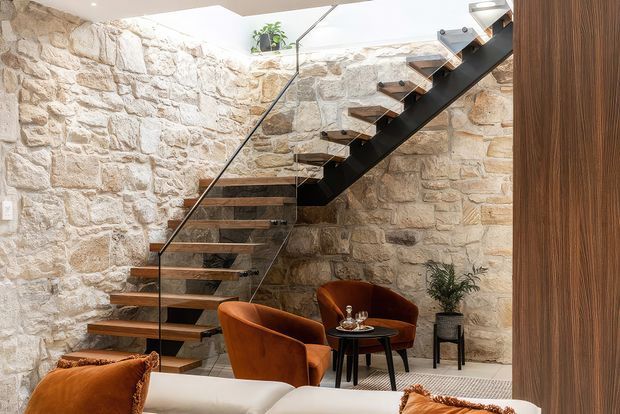 A refined interior featuring a floating staircase with dark metal stringers, timber treads, and a clear glass balustrade set against exposed pale stone walls. A small lounge seating area with rust-toned upholstered chairs, a round black table, plants, and a partial sofa occupies the space beneath and beside the stairs. A large roof or ceiling opening brings in abundant natural light.