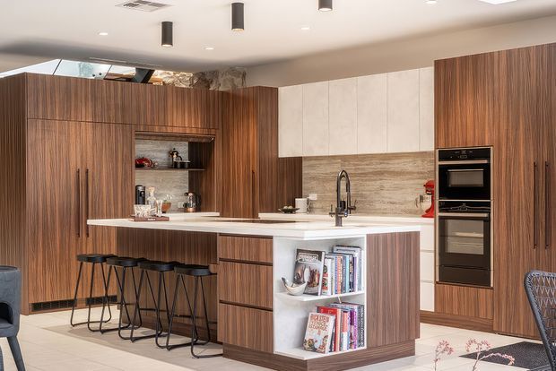 Contemporary kitchen with extensive dark timber-grain cabinetry, a white island with integrated open shelving, white benchtops, a textured stone-look splashback, and a built-in black oven tower. The island incorporates seating for four with slim black bar stools. Recessed and pendant-style ceiling lighting create a refined, warm interior.