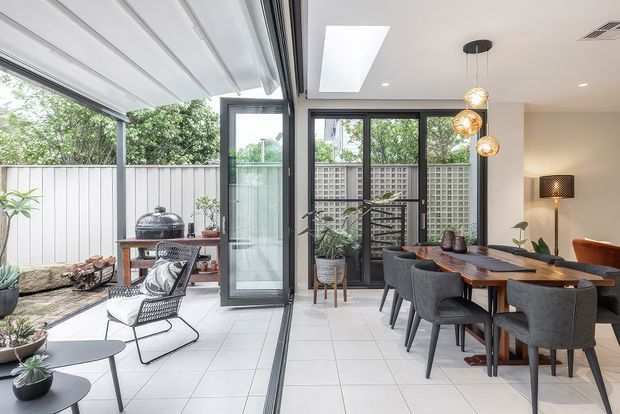 Bright contemporary dining and indoor-outdoor living area with a tiled floor, large black-framed sliding and folding glass doors, a covered patio, and a dining table with upholstered chairs. The room includes pendant globe lighting, a skylight, indoor plants, and views toward a screened courtyard or garden.