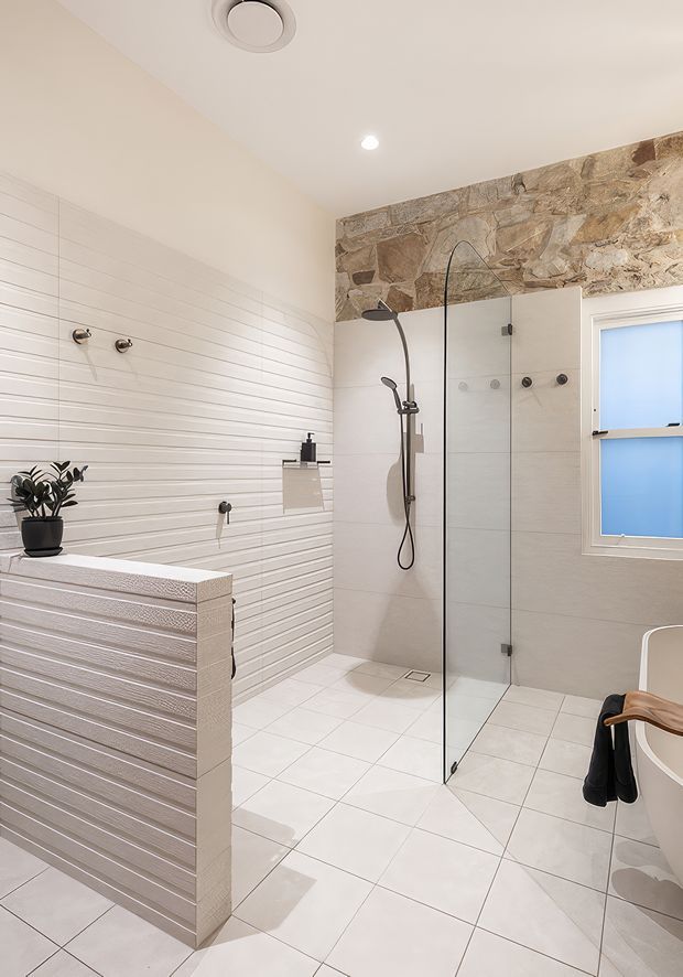 Bright contemporary bathroom featuring a large open shower enclosed by frameless glass, white tiled walls and floor, a textured horizontal wall tile feature, and a natural stone accent wall above the shower. A freestanding bathtub is partially visible at right, with a small tiled partition and potted plant in the foreground.