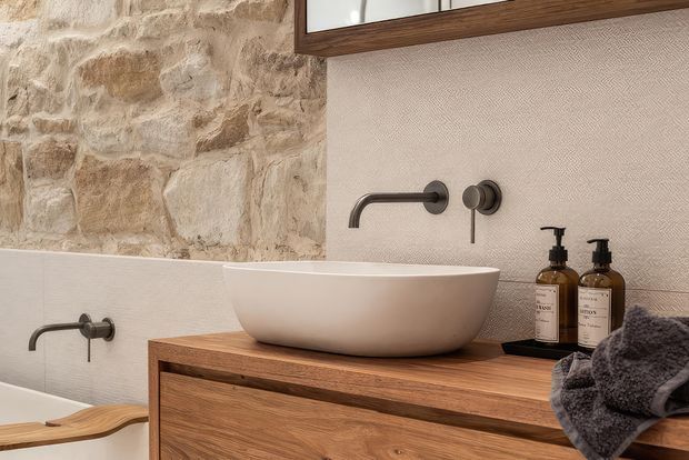 Close-up view of a warm, textural bathroom vanity with a white vessel basin, dark wall-mounted tapware, timber cabinetry, and a partially visible bathtub. The walls combine rough natural stone with subtle textured plaster, creating a refined rustic feel.