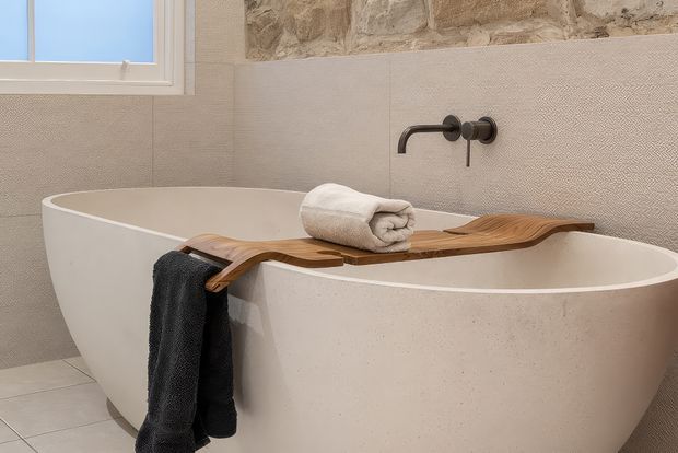 A serene bathroom featuring a freestanding white oval bathtub beneath a wall-mounted dark metal tap. A sculptural timber bath caddy spans the tub, holding neatly rolled towels, while a dark towel hangs over the side. The room has warm textured beige wall tiles, a natural stone feature section, pale floor tiles, and a small frosted window.