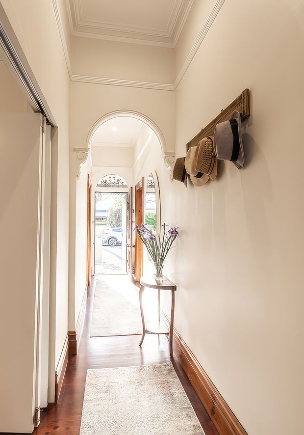 A narrow, elegant residential entry hallway with warm polished timber flooring, cream-painted walls, ornate layered ceiling cornices, decorative skirting boards, and a carved arched opening leading to a glazed front door. A small round console table with flowers and a wall-mounted hat rack add detail, while a runner defines the circulation space. The overall character is bright, refined, and traditionally styled.