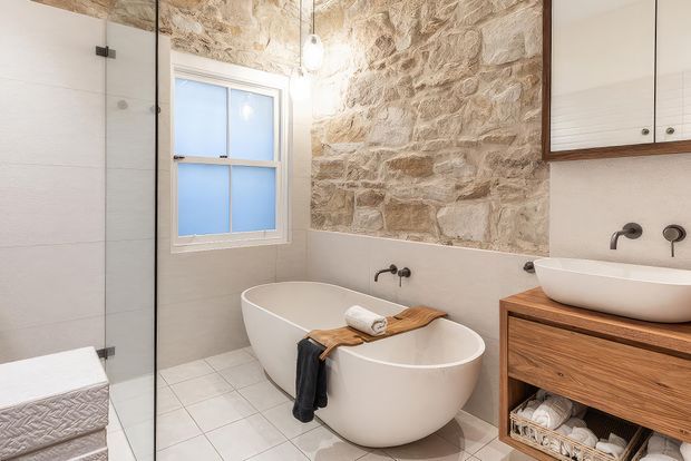 A bright, refined bathroom featuring a freestanding white bathtub beneath a rustic exposed stone feature wall, a frameless glass shower enclosure, pale tiled walls and floor, and a timber vanity with a white vessel basin. A window provides natural light, while wall-mounted black fixtures and a hanging pendant add contrast.