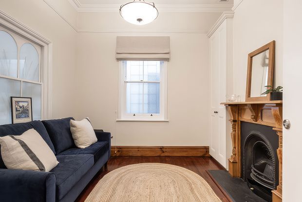 A compact, refined living room with a deep navy sofa, cream cushions, a natural woven oval rug, polished dark timber flooring, and a traditional timber fireplace with black cast-iron detailing. The room features ornate ceiling cornices, decorative wall mouldings, a sash window with a Roman blind, a built-in arched glazed cabinet or pass-through on the left, and a large timber-framed mirror above the mantel. The palette is warm and neutral with classic architectural detailing.