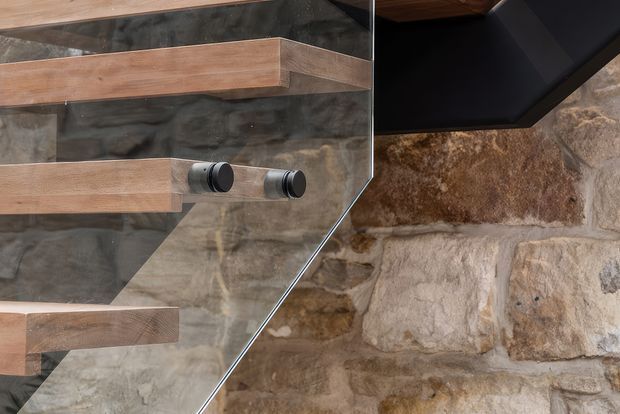 Close-up architectural detail of a contemporary stairway, showing thick timber stair treads and a clear glass balustrade with dark metal cylindrical fixings. A rough natural stone wall forms the backdrop, with dark timber or painted ceiling elements visible above.