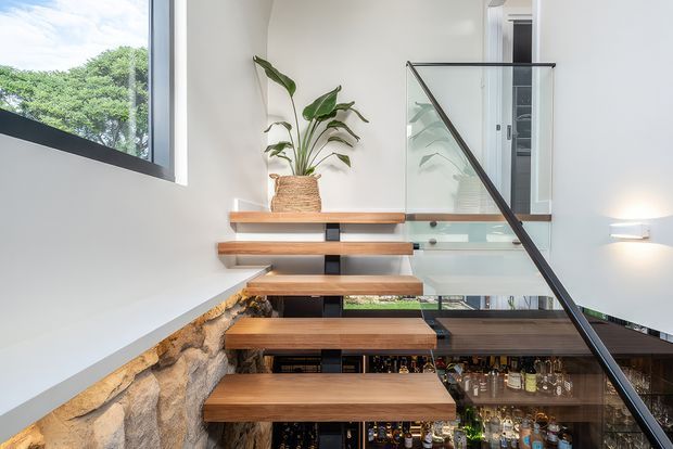 Modern interior stairway with floating timber treads, a black metal structural stringer, and clear glass balustrading. The stair descends beside a rough natural-stone wall into a visible glass-fronted bottle storage or cellar area, while a potted plant sits on an upper landing. Large glazing brings in bright natural light.