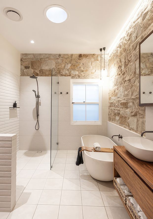Bright contemporary bathroom featuring a freestanding white bathtub, wall-mounted vessel basin, frameless glass shower enclosure, large-format pale floor and wall tiles, and a rustic natural-stone feature wall. A timber vanity with open storage runs along the right side, with a large mirror and recessed ceiling lighting.