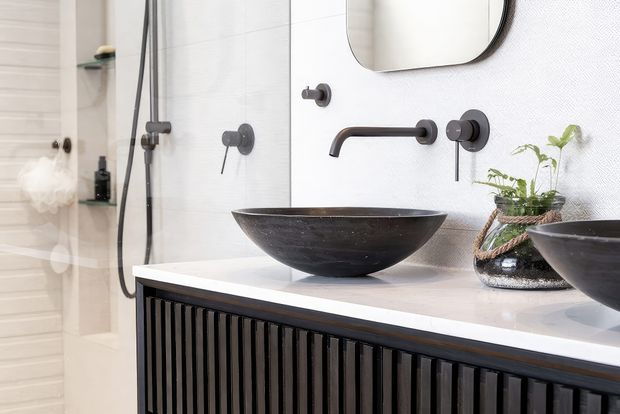 Contemporary bathroom vanity featuring a dark fluted timber cabinet, white countertop, matte black wall-mounted tapware, and two black vessel basins. A frameless shower with matching dark fixtures, rounded mirror, and potted greenery complete the refined monochrome space.