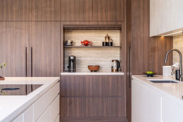 Contemporary kitchen cabinetry with a warm dark timber veneer finish, white stone benchtops, integrated open shelving, and built-in coffee and tea storage. A sink with a high-arch mixer tap is visible on the right, while the left side includes additional cabinetry and a partially visible island or peninsula. The open shelves display small appliances and serving ware.