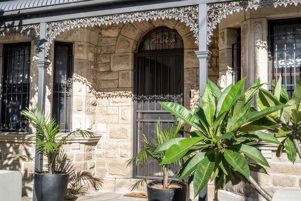 A sunlit heritage-style stone facade with arched openings, barred windows and door, ornate white-painted lacework trim, and corrugated metal roofing. Tropical potted plants frame the entrance and soften the masonry exterior.