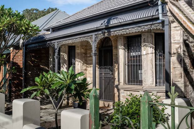 Front exterior of a heritage terrace or cottage featuring ornate cast-iron lacework, sandstone masonry, barred timber-framed windows and a dark corrugated metal verandah roof. A small landscaped front garden and decorative metal fence sit in front of the facade.
