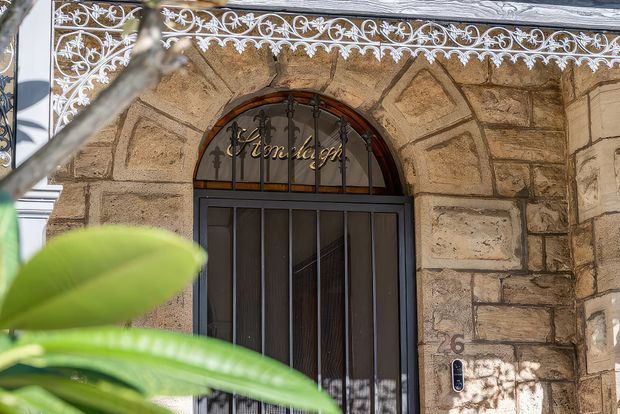 Close-up exterior view of an ornate arched entry door set within a rough-cut sandstone masonry facade. The doorway has dark metal-framed glazing and security bars, decorative wrought-iron trim and signage above, and a white-painted ornamental metal balcony or parapet railing across the top. Tropical foliage partially obscures the foreground.