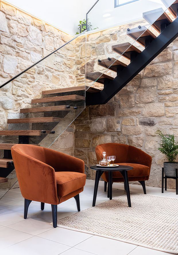 A bright, refined interior featuring a sculptural open-riser staircase with dark structural stringers, warm timber treads, and clear glass balustrades. Rough natural stone walls surround a small lounge area furnished with two rust-orange upholstered armchairs, a black side table, and a neutral woven rug. A skylight or high-level glazing brings in abundant natural light, with potted greenery adding a subtle organic touch.