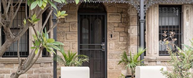 Close view of a traditional stone residential facade with a dark timber or metal entry door, barred windows, ornate lacework verandah detailing, black posts and trim, and potted tropical greenery.
