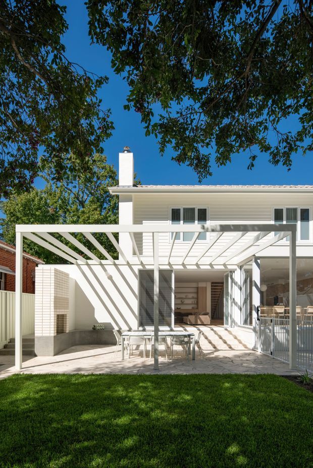 Bright rear exterior view of a contemporary two-storey home with a white rendered facade, flat-looking low-pitched roof, chimney, and large glazed openings. A white pergola shades a paved outdoor dining terrace, with a built-in outdoor fireplace against a pale brick wall and a covered patio/lounge area opening directly into the house. Mature trees and lawn surround the backyard.