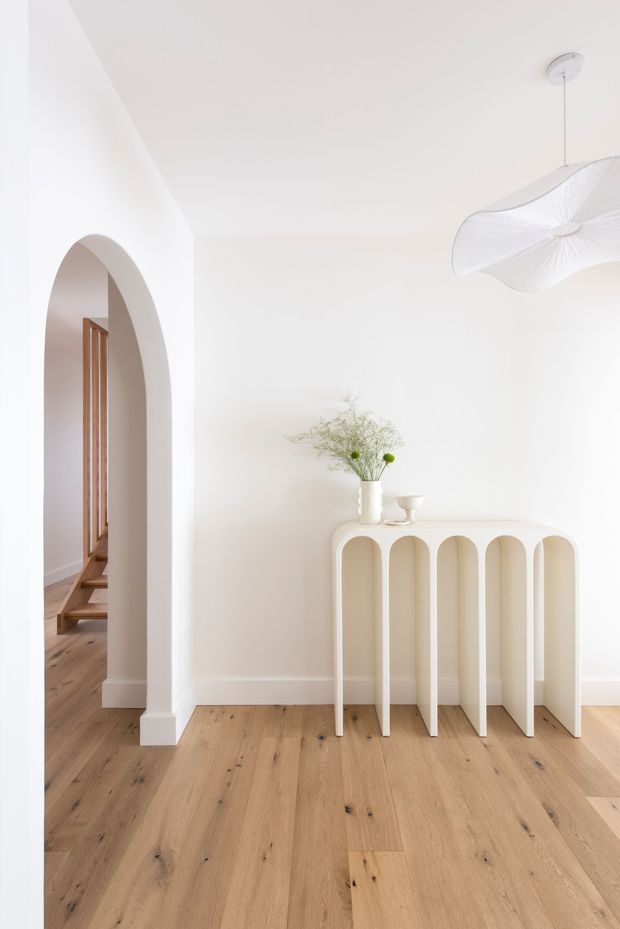 Bright minimalist residential hallway or entry space with white walls, a rounded arched opening leading to a timber staircase, pale natural timber flooring, and a sculptural cream console table with repeated arched legs. A delicate white pendant light and simple floral arrangement add soft decorative detail.