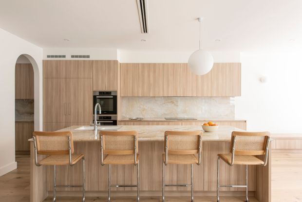 Bright, spacious contemporary kitchen with a large light-wood island and four woven cane bar stools. Full-height light timber cabinetry, integrated appliances, a pale stone-look splashback and benchtops, white walls and ceiling, timber flooring, and a spherical pendant light create a warm minimalist interior. An arched opening leads to pantry or storage joinery at the left.