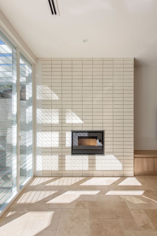 Bright, minimalist interior featuring a modern built-in fireplace set into a full-height wall of small rectangular cream tiles. Large louvred glazing on the left floods the room with sunlight and casts strong linear shadows across the tiled floor and walls. A low timber built-in cabinet runs along the right wall, and the floor is finished in large warm beige stone or porcelain tiles.