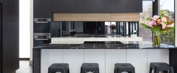 Contemporary kitchen featuring a large island with seating for four, white cabinetry, a dark stone benchtop, tall matte-black joinery, timber overhead cabinetry, integrated ovens, and a mirrored splashback. Large windows provide abundant natural light and views of greenery outside.