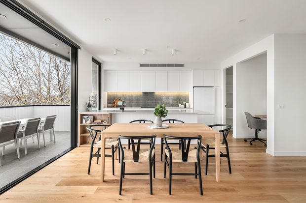 Bright open-plan kitchen and dining area with white flat-panel cabinetry, a tiled backsplash, central island, timber flooring, and a large timber dining table with black-framed chairs. Floor-to-ceiling glazed sliding doors open to an outdoor dining area, while a study nook is visible through the opening on the right.