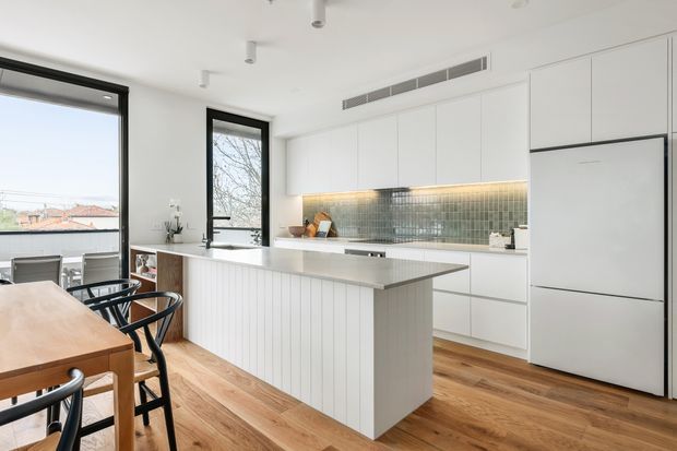 Bright, contemporary open-plan kitchen and dining area with extensive white flat-panel cabinetry, a large central island, integrated white refrigerator, and a muted green-grey tiled splashback illuminated by under-cabinet lighting. Warm timber flooring and a timber dining table contrast with black-framed windows and black dining chairs. The space has generous natural light and views to neighbouring rooftops.