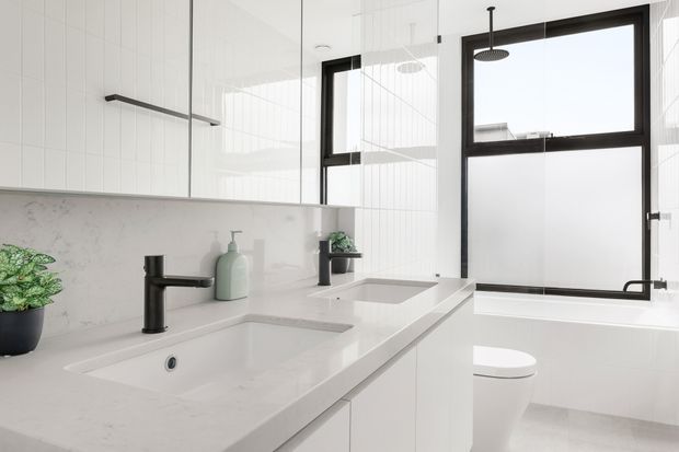 Bright contemporary bathroom featuring a double vanity with integrated white basins, pale stone surfaces, white vertical wall tiling, frameless mirrored cabinetry, matte black tapware and shower fixtures, and a freestanding white bathtub beside black-framed windows. A toilet and understated greenery complete the clean, minimal space.