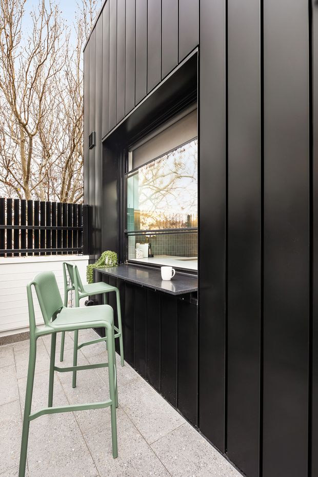 A compact outdoor terrace or courtyard nook beside a contemporary building, featuring black vertical metal cladding, a built-in servery or window counter with dark benchtop, pale terrazzo-style paving, and two sage-green bar stools. A black slatted privacy fence and leafless trees are visible beyond the space.