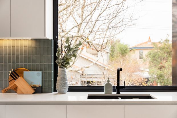 Bright contemporary kitchen interior featuring pale flat-panel cabinetry, a dark green-gray vertical tile splashback, a white benchtop, and a black undermount sink with matching gooseneck tap. A large window provides abundant natural light and overlooks neighboring homes and trees. A timber knife block and ceramic vase add warmth to the streamlined fit-out.