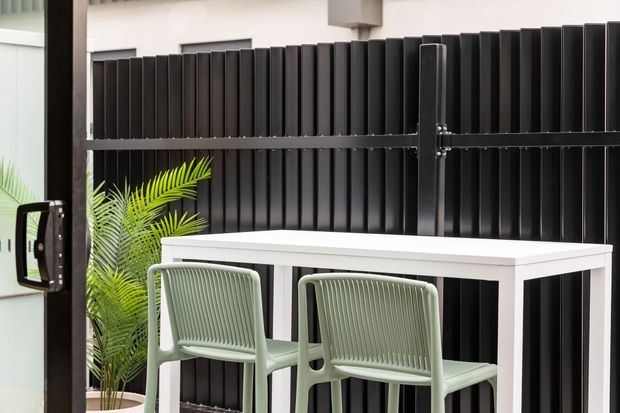 A compact outdoor entertaining area with a white bar-height table and two pale green woven chairs positioned against a tall black vertical-slat privacy fence. A potted palm adds greenery, while a dark-framed glass door or window is visible at the left edge.
