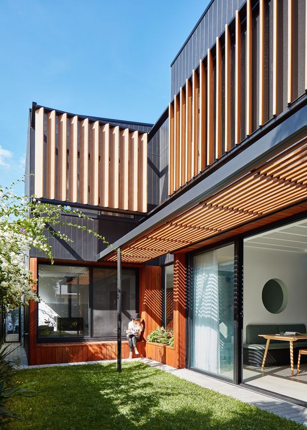 A contemporary multi-level residential courtyard viewed from a small landscaped lawn. The building combines dark vertical metal cladding with warm timber slatted screens and timber-lined soffits. Large glazed sliding doors open to an interior dining or living area, while projecting upper volumes feature repeated vertical timber fins for sun control and visual rhythm. Strong sunlight creates patterned shadows across the facade and courtyard.