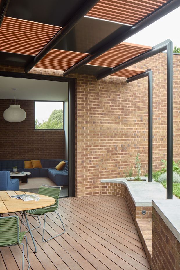 A covered outdoor entertaining deck beside a brick-walled home, featuring a timber deck, black steel pergola structure with horizontal timber slats, outdoor dining furniture, and an adjoining indoor living area visible through wide open glazing. A raised concrete-edged garden bed and brick steps border the space.