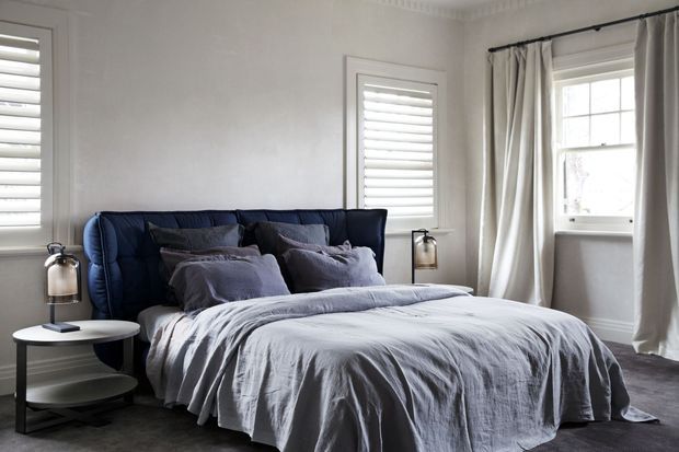 A refined bedroom with a deep navy upholstered bed, layered grey-blue bedding and pillows, and white walls with decorative cornice detailing. Plantation shutters cover the narrow windows, while full-length pale curtains frame a larger sash-style window. Dark flooring, minimalist round bedside tables, and matching lantern-style lamps create a calm, sophisticated atmosphere.