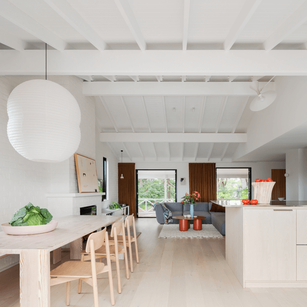 Bright open-plan interior combining a kitchen, dining area and living room. The space features white-painted exposed ceiling beams and rafters, white brick walls, pale timber flooring, a light timber dining table with matching chairs, a large white pendant light, a white kitchen island, and timber slatted curtains framing views to greenery outside.