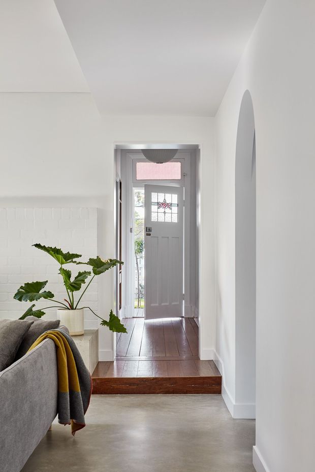A bright residential entry hall viewed from an adjoining living area. The space features white walls, a white-painted brick feature wall, a grey front door with decorative glazed panels, dark stained timber floorboards at the entry landing, a polished concrete floor in the foreground, and a curved wall opening. A grey sofa with a mustard and charcoal throw and a large leafy indoor plant are partially visible.
