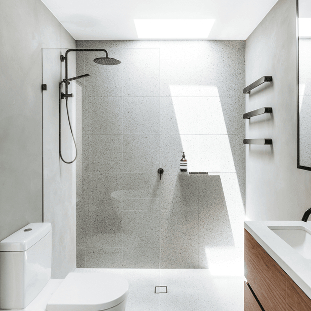 Bright contemporary bathroom featuring a walk-in shower with dark metal fixtures, large light-speckled wall and floor tiles, a white toilet, and a timber-fronted vanity with white basin. Strong daylight enters from a high opening or skylight, creating a clean, minimalist atmosphere.