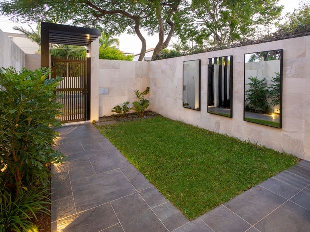 A landscaped courtyard or entry garden with pale stone-clad boundary walls, a dark tiled pedestrian path, rectangular lawn, integrated planting, and a black timber or metal slatted entry gate beneath a modern pergola-like canopy. Three tall black-framed mirrored or glazed wall openings reflect the garden and gate, adding depth. Mature trees and dense greenery create a private, contemporary outdoor arrival space.