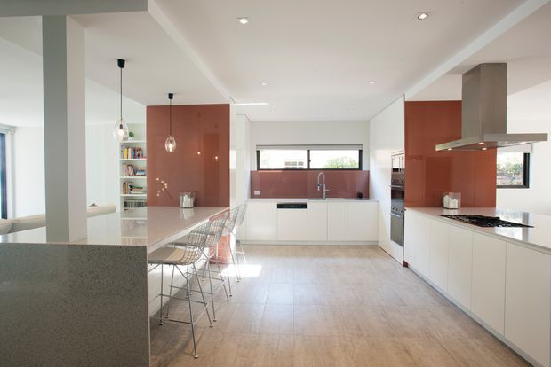 Bright, spacious contemporary kitchen with white flat-panel cabinetry, a long island with transparent bar stools, and a pale stone benchtop. Glossy terracotta-red splashback panels and matching wall cabinetry add colour, while a stainless-steel rangehood and integrated appliances line the cooking zone. Pale timber-look flooring, recessed ceiling lights, pendant lights, and broad glazing create an airy open-plan space.