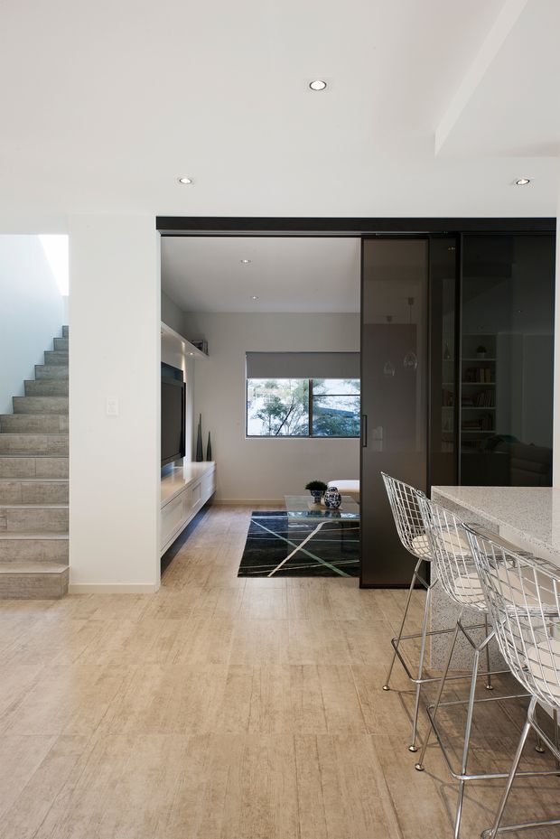 Bright contemporary open-plan interior showing a kitchen and living area. The foreground features a pale stone or engineered-stone island with white wire-frame bar stools, while a dark glazed sliding partition and built-in shelving separate the living room. Pale large-format floor tiles, white walls, recessed ceiling lights, a staircase with tiled treads, and black-and-white geometric furnishings create a clean modern composition.