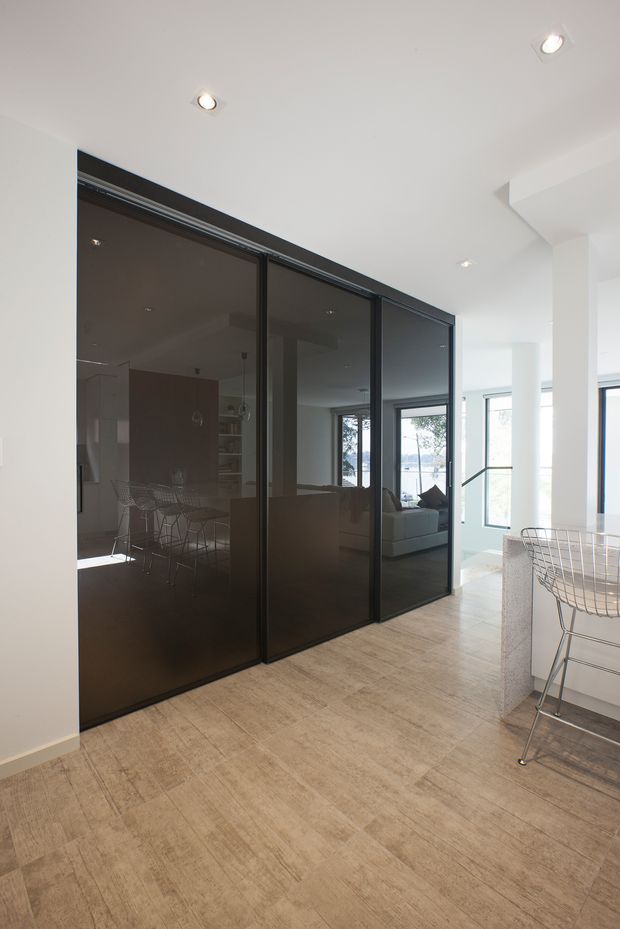Bright contemporary interior showing a circulation area with large dark-framed glazed sliding partitions enclosing a dining or living space. The room has pale timber-look flooring, white walls and ceiling with recessed downlights, and a light stone or solid-surface peninsula with wire-frame bar stools. A living area and dining furniture are visible through the glazing, with abundant natural light from full-height windows.