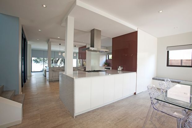 Open-plan contemporary kitchen and dining area with a large white island, integrated cooktop and stainless-steel rangehood, glossy dark red-brown cabinetry, pale timber-look flooring, and a glass dining table with transparent chairs. The space is bright and minimalist, with a hallway and living area extending beyond the kitchen.