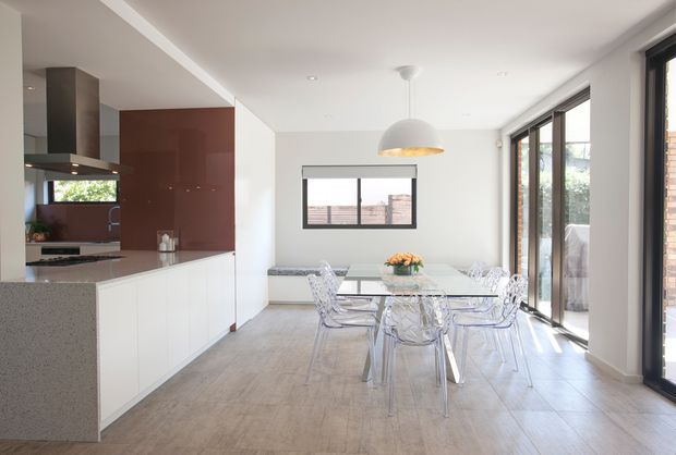 Bright open-plan kitchen and dining area with white flat-panel cabinetry, a dark reddish-brown feature splashback or wall panel, pale stone benchtops, timber-look flooring, a glass dining table with transparent acrylic chairs, and extensive black-framed glazing along one side. A contemporary pendant light hangs above the dining table.