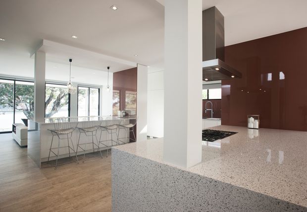 Bright open-plan kitchen and living area with a large speckled stone kitchen island and peninsula, glossy deep burgundy splashback cabinetry, white walls and ceilings, timber flooring, pendant lights, and extensive floor-to-ceiling glazing. Wire-frame bar stools line the island, while a living area is visible beyond.