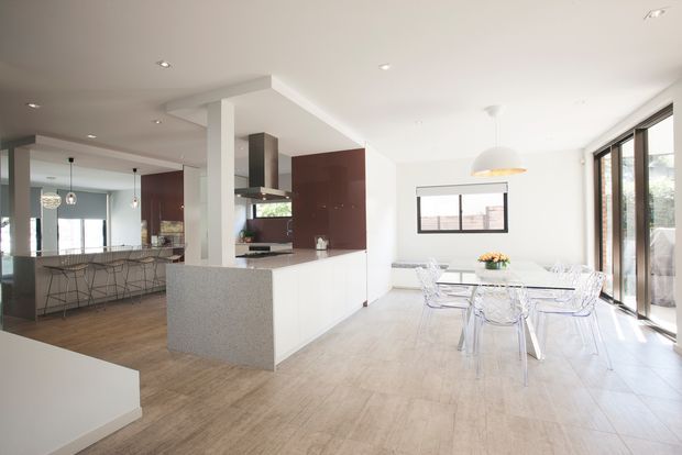 Bright open-plan contemporary kitchen and dining area with a large white island, integrated stainless-steel rangehood, dark wood cabinetry, pale timber-look flooring, and a transparent dining table with clear chairs. Floor-to-ceiling glazed doors and multiple windows bring in abundant natural light.