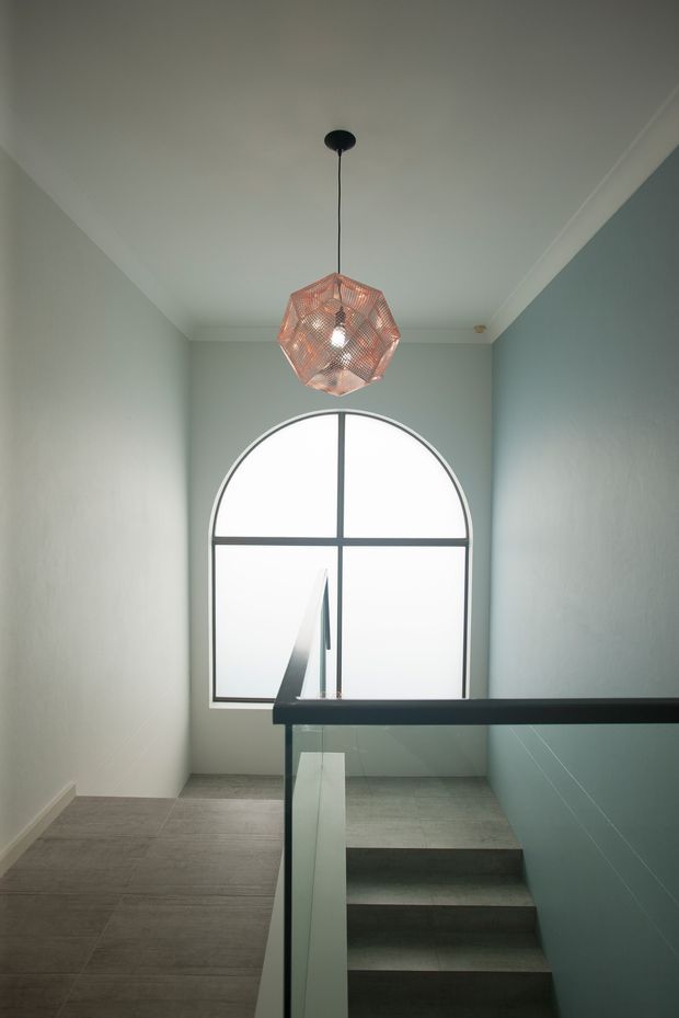 A modern stair landing with grey tiled flooring, pale blue-grey walls, crisp white cornices, and a large arched window with dark framing. A sculptural woven pendant light hangs from the ceiling above the stairwell, while a glass balustrade and dark handrail overlook the stairs. The space is softly lit and restrained in palette, with a clean contemporary feel.