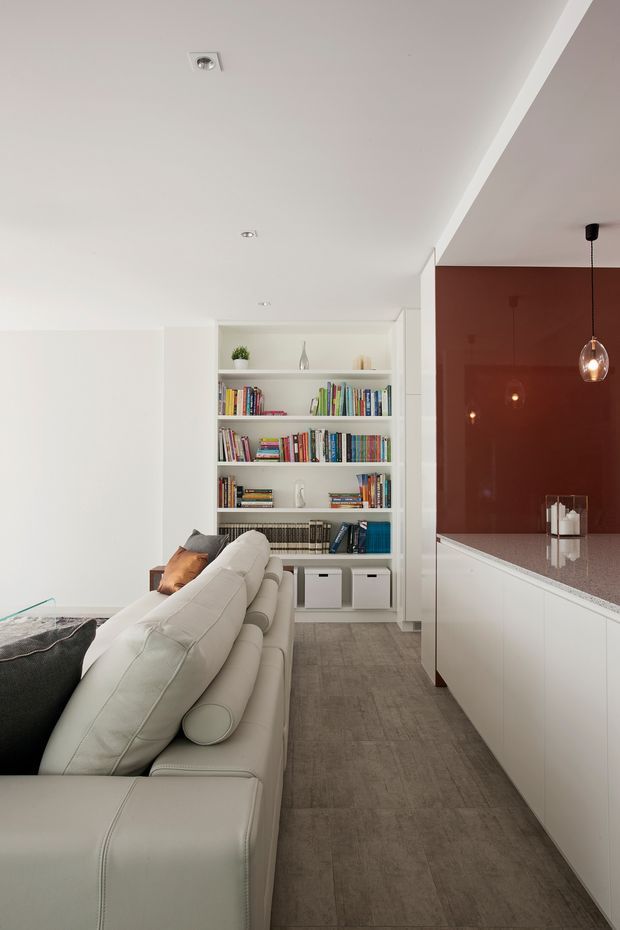 A contemporary open-plan living area viewed toward a built-in white bookshelf and adjacent kitchen. A large pale sectional sofa occupies the foreground, with grey-brown tiled flooring, white cabinetry, a speckled light benchtop, recessed ceiling lights, and a deep terracotta-red feature wall behind a hanging pendant light.